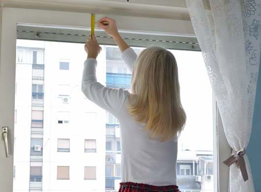 Blonde woman measuring and marking a position of a hole in order to install a blind. Residential building is in a background